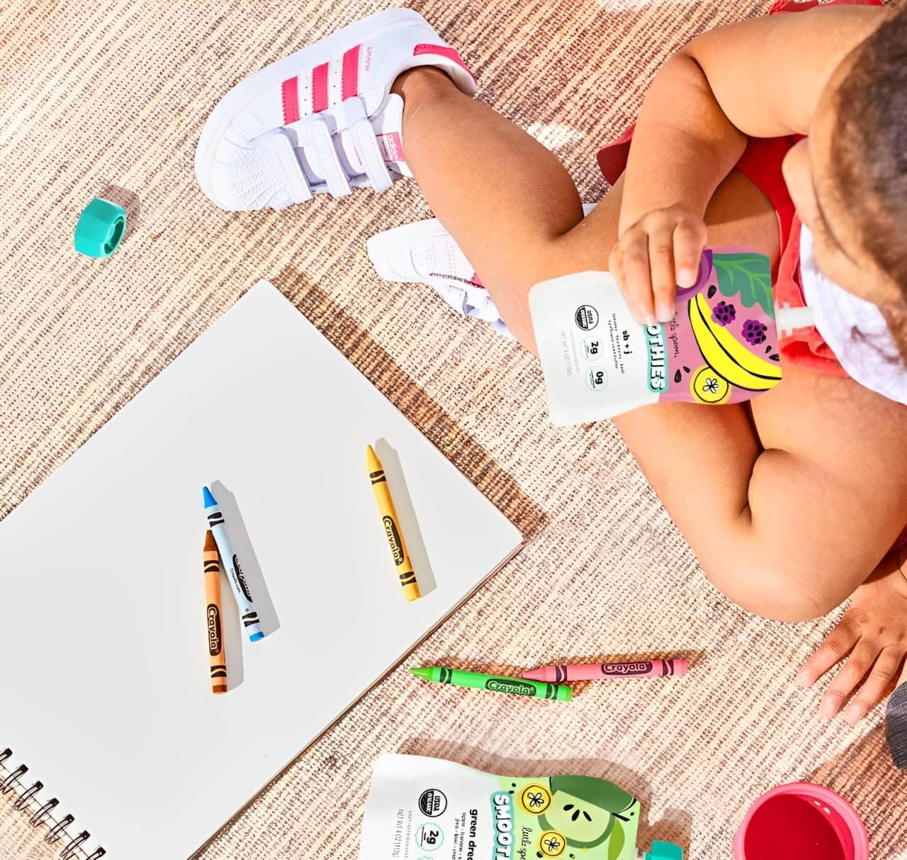 Baby sitting with crayons and a notebook while drinking a Little Spoon Smoothie pouch