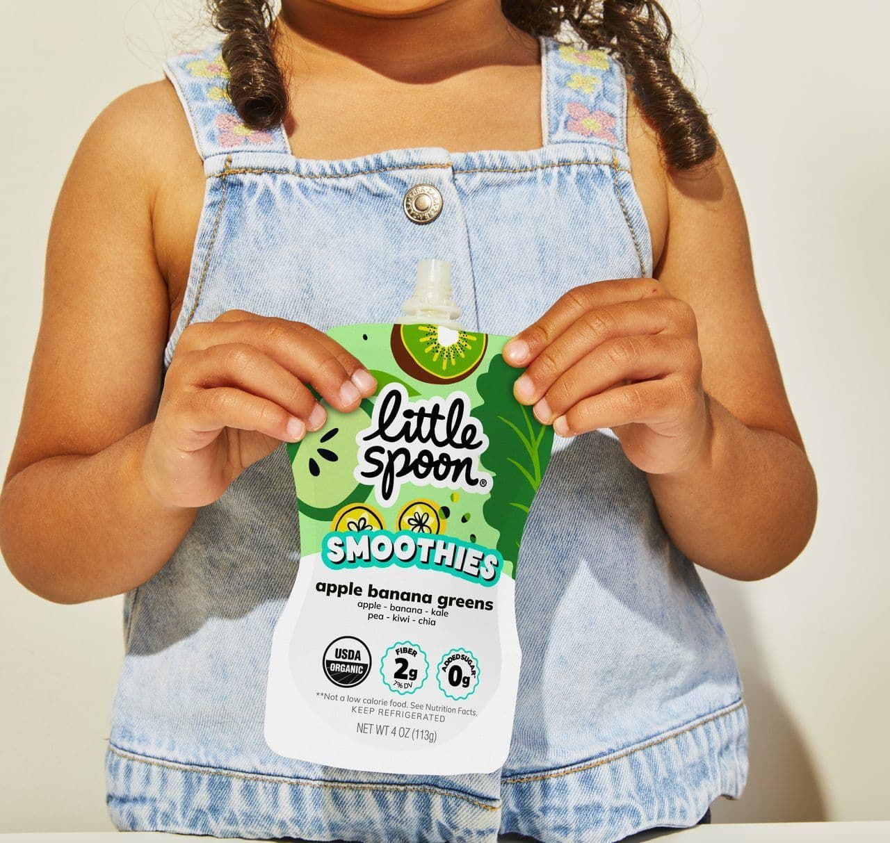 Child in denim overalls holding a Little Spoon Apple Banana Greens Smoothie pouch