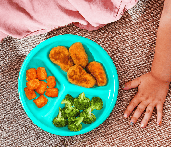 A toddler's hand sits next to a teal-colored plate of chicken nuggets, broccoli and sweet potato bites.