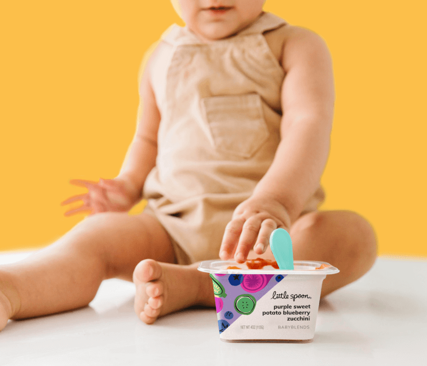 The bottom half of a young toddler sits with a Little Spoon babyblend mixture of purple sweet potato, blueberry and zucchini. A teal spoon is seen sticking out of the puree next to some tiny toes.